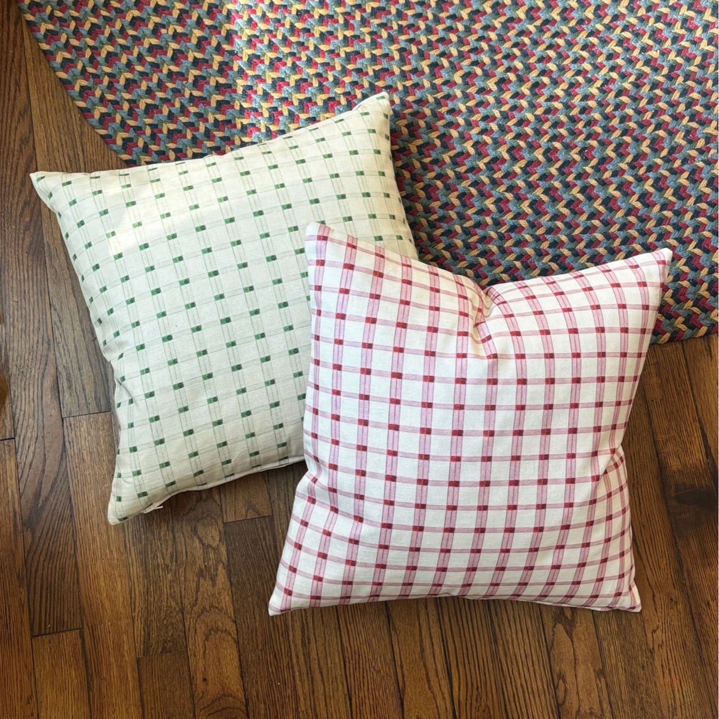 Two square throw pillows with gingham patterns, one in green on a white background and the other in red on a white background, styled on a wooden floor in front of a colorful woven rug.
