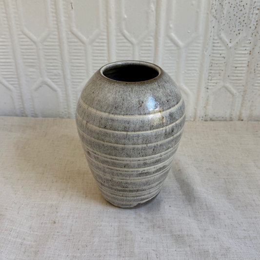 A small ceramic vase with a rounded bulbous shape, featuring horizontal ribbed ridges and a speckled gray-brown glaze, set on a light beige fabric surface against a textured white background.