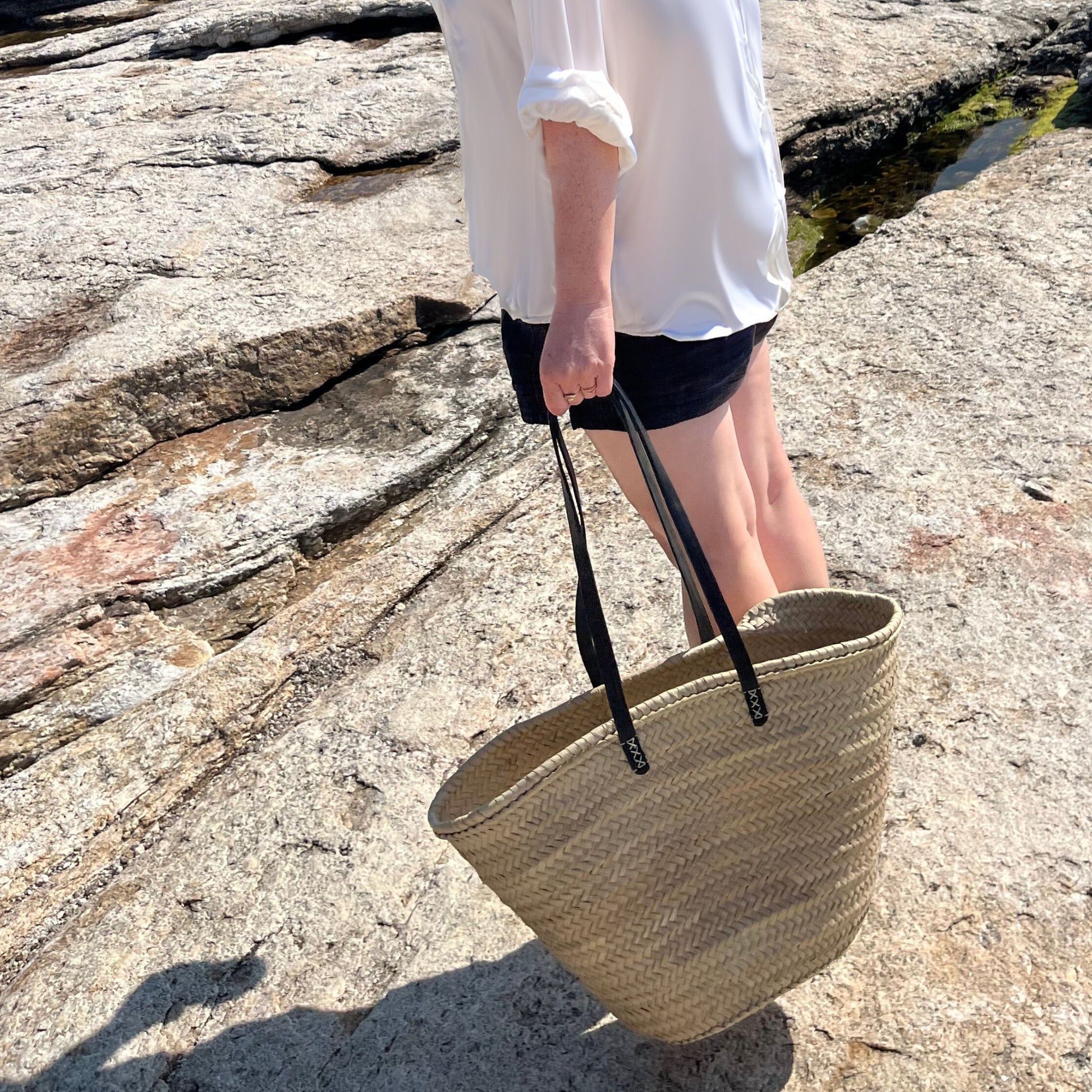 French market tote with black leather shoulder straps being held by someone outside standing on rocks.