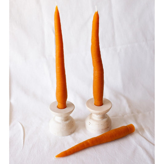 Two carrot-shaped beeswax candles on white candle holders, with a single carrot candle beside them, all displayed on a white fabric surface.