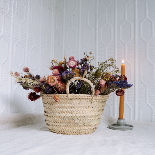 Woven basket filled with dried flowers on a white surface with a candle in the background.