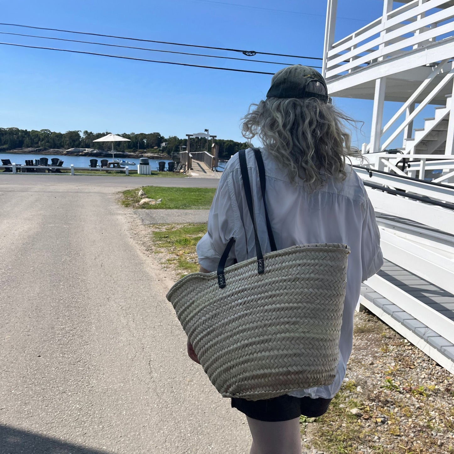 Woman holding french market tote with black leather shoulder straps next to a white house on her way to the beach in the background.