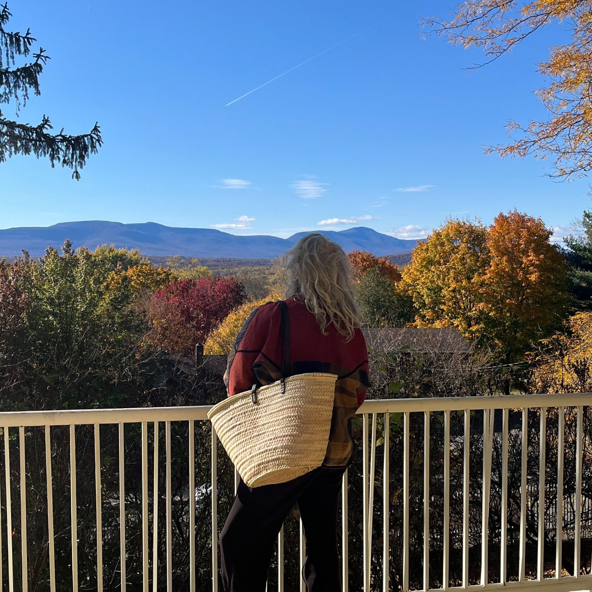 French market tote with black leather shoulder straps being worn by a woman on a porch overlooking the fall colored woods and the mountain range.