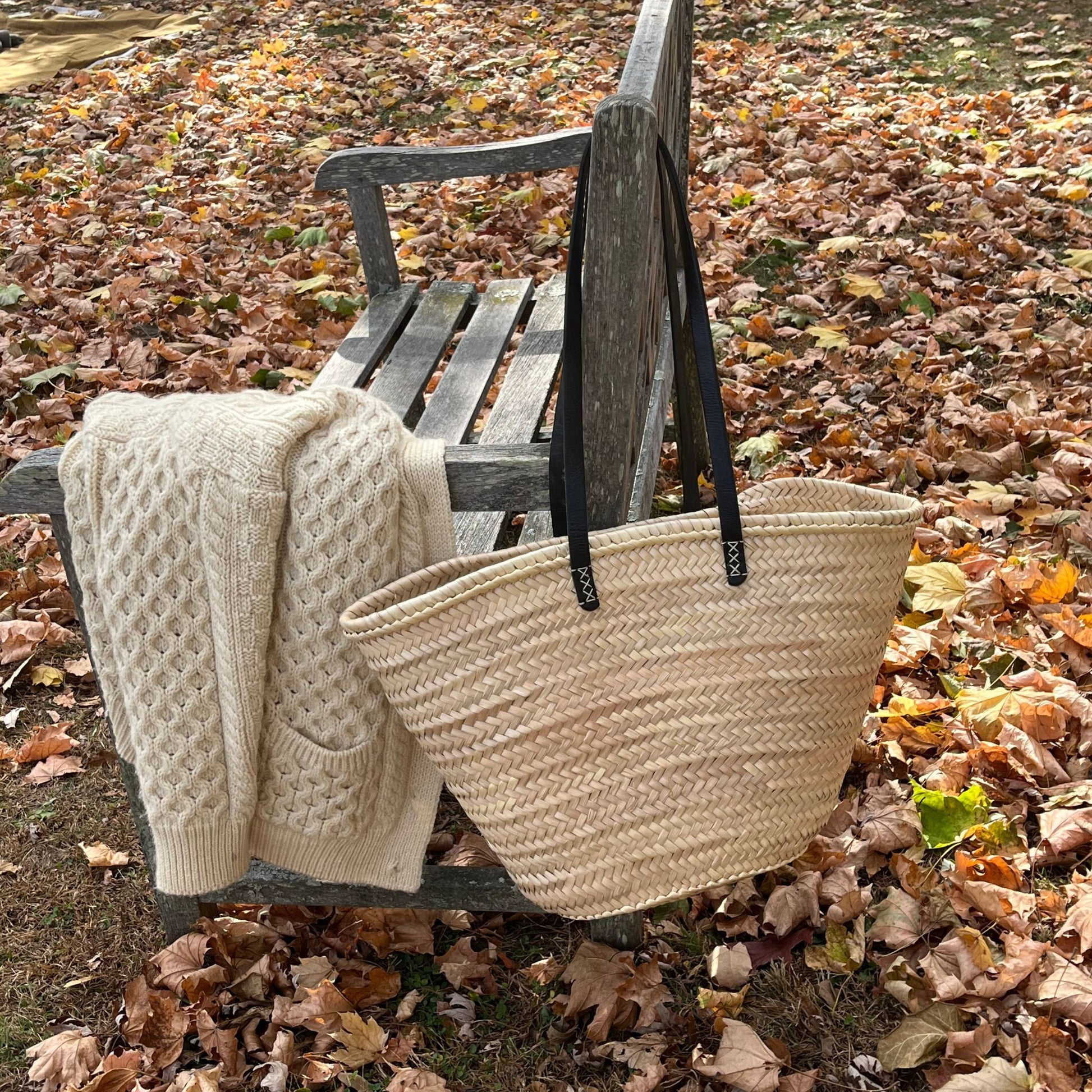 French market tote with black leather shoulder straps hanging from an outside bench with a white sweater draped along the arm of the seat and fall leaves in the background.