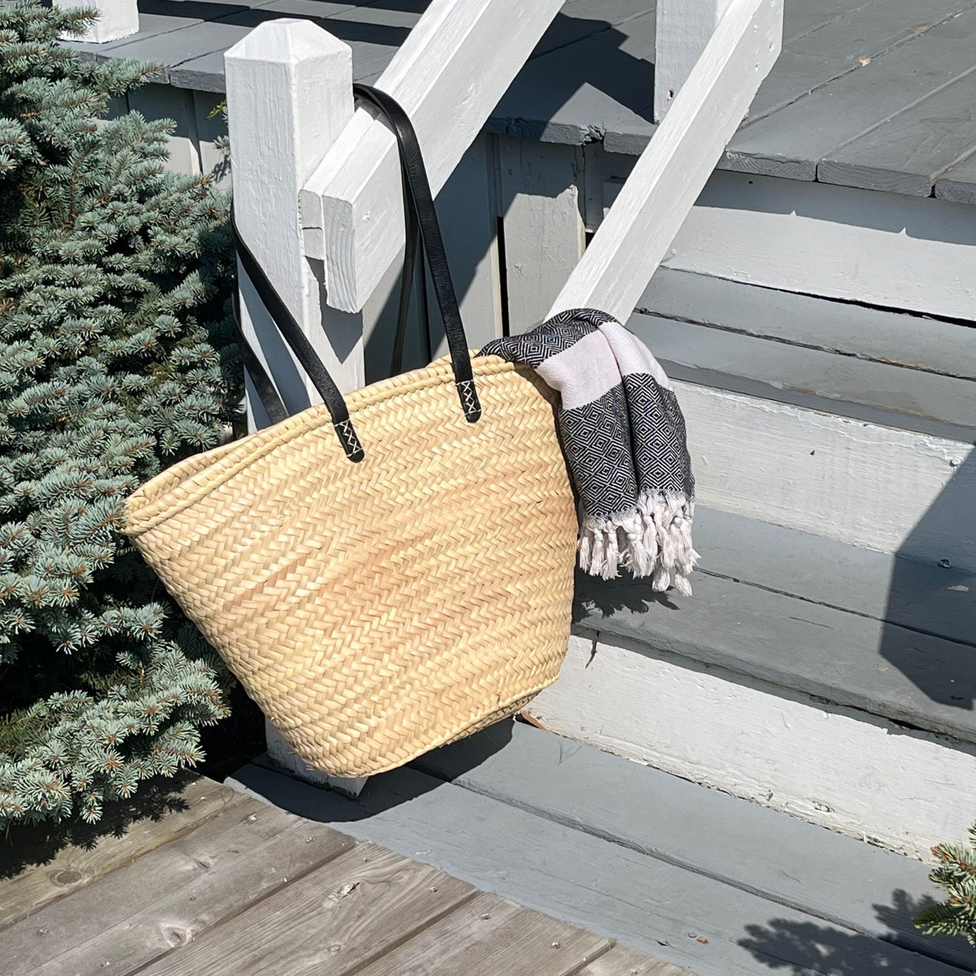 French market tote with black leather shoulder straps hanging from a porch railing with a blanket hanging from it and bushes on either side.