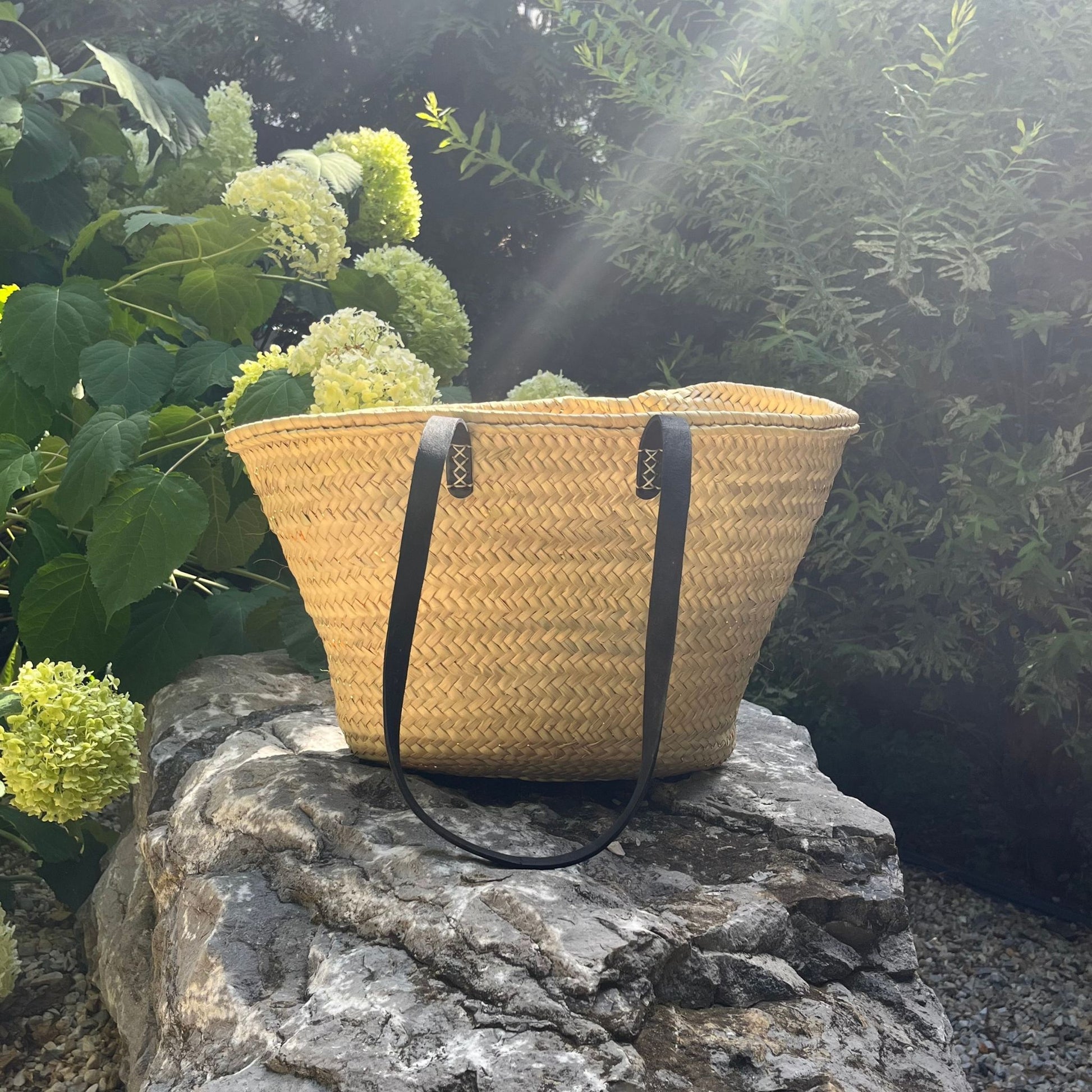 French market tote with black leather shoulder straps on a rock with hydrangeas and greenery in the background and sunlight from the top right corner.