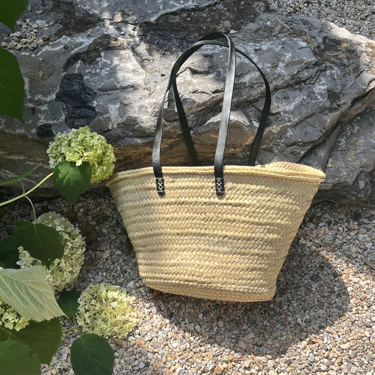 French market tote with black leather shoulder straps leaning against a rock with hydrangeas alongside it.