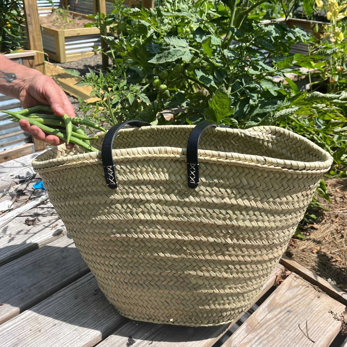 French market tote with black leather shoulder straps outside on a porch with greenery in the background and a hand putting vegetables inside.