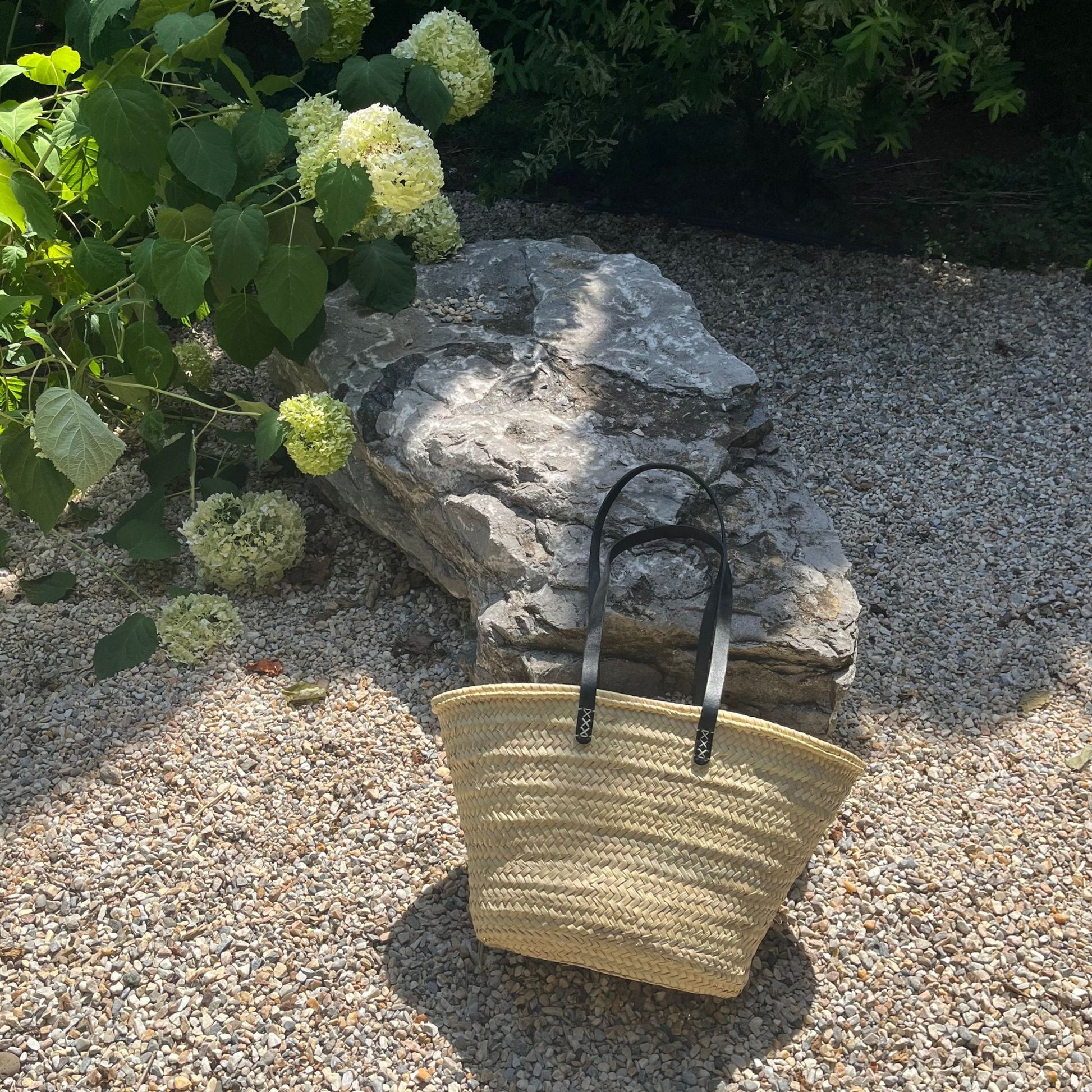 French market tote with black leather shoulder straps leaning against a rock with hydrangeas and greenery in the background.