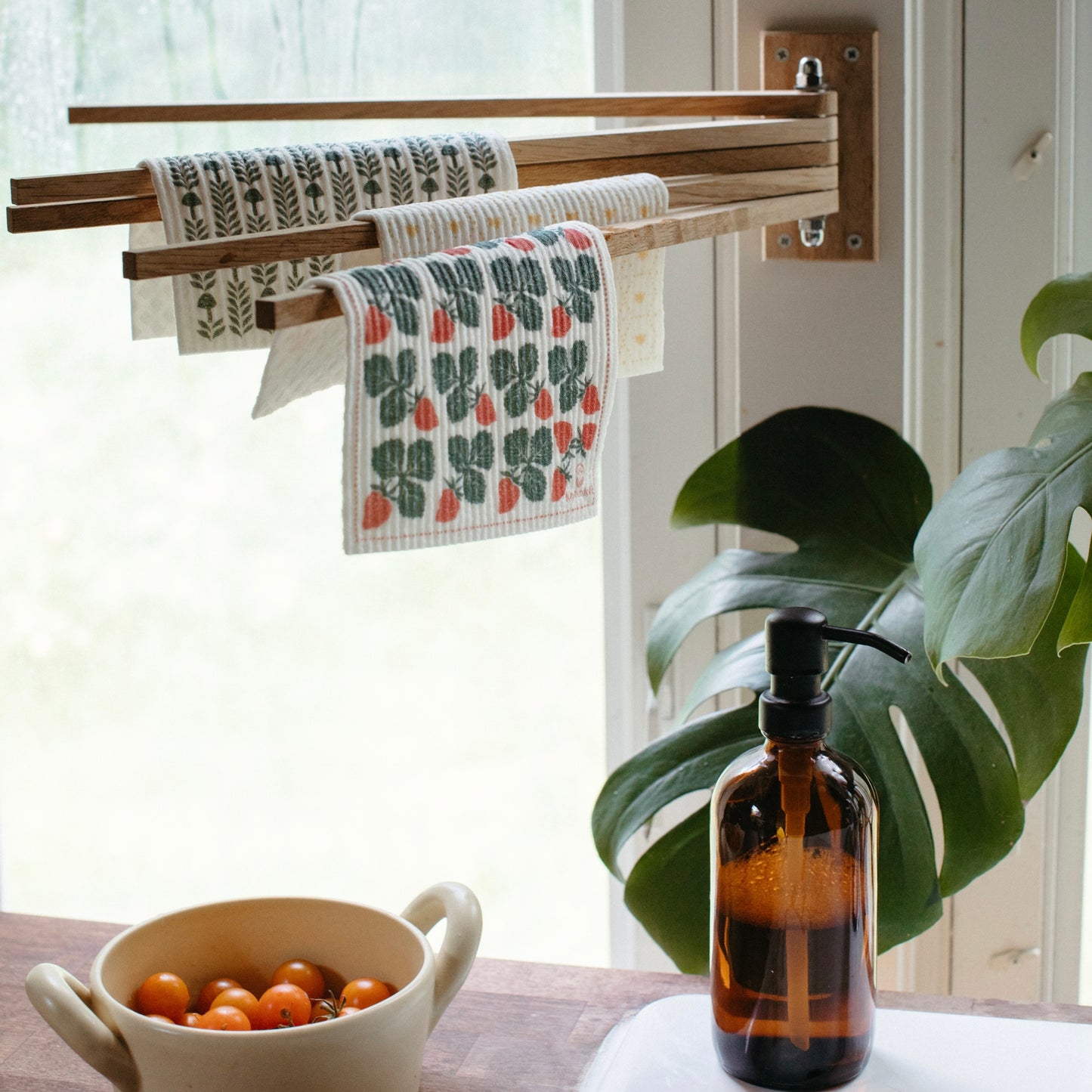 Oak wood towel drying rack holding Swedish Dishcloths next to a soap jar and a bowl of tomatoes.