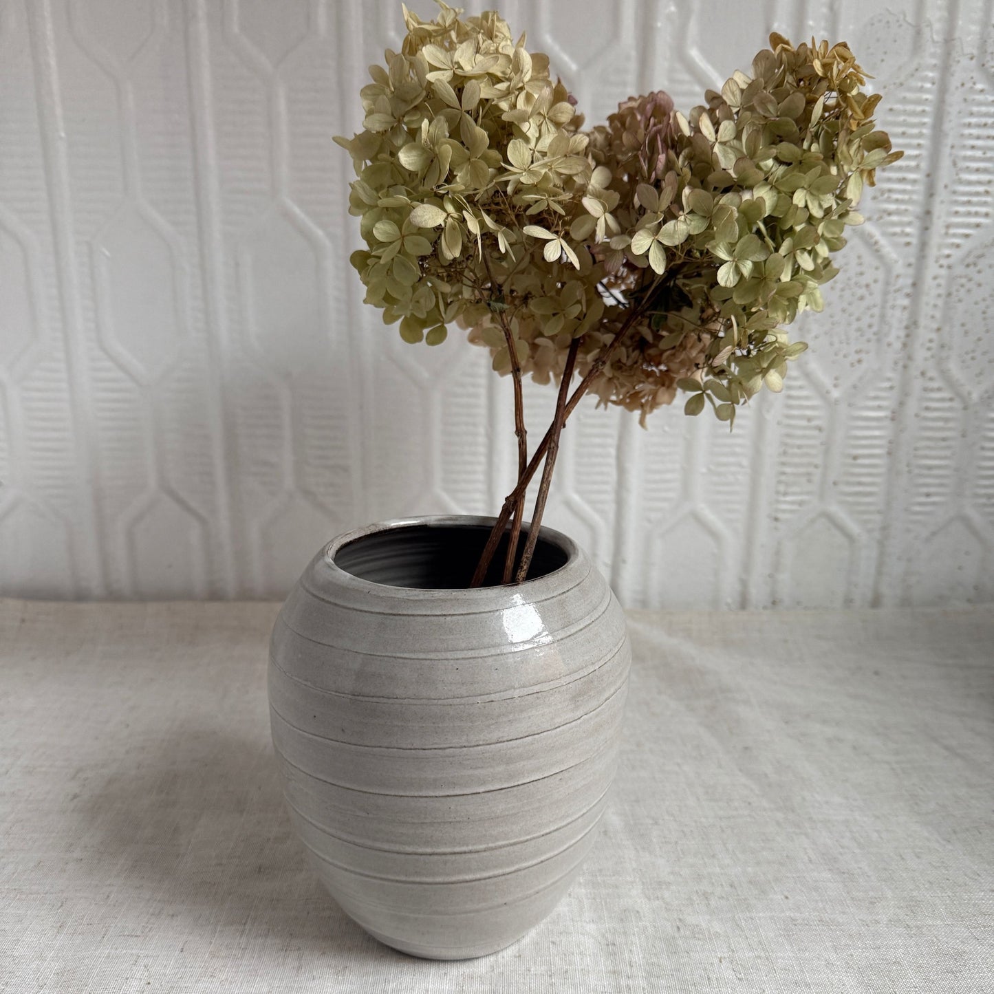 Gray ceramic vase with dried flowers against a textured white wall.