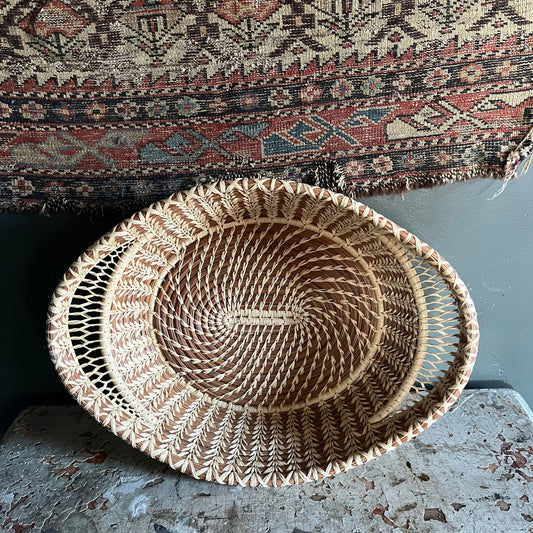 A tan and brown pine needle basket with intricate lace-like raffia handles. In the background is a rug.
