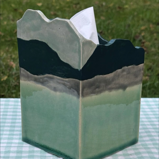 A ceramic tissue box cover shaped like layered mountains in sage green, gray, and dark green, with a white tissue inserted, set on a green-and-white checkered tablecloth with grass in the background.
