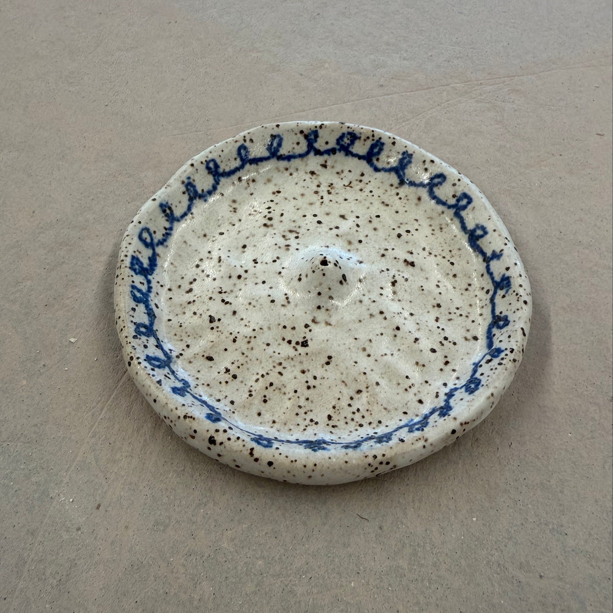 A handmade ceramic dish with a textured, speckled off-white glaze and a blue decorative border, photographed on a neutral gray surface.
