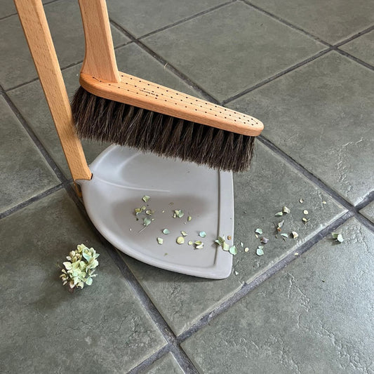 A standing broom and dustpan set cleaning flowers off of a tiled floor.