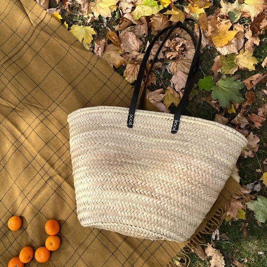 French market tote with black leather shoulder straps laying on a brown picnic blanket on top of fall foliage with oranges alongside it.