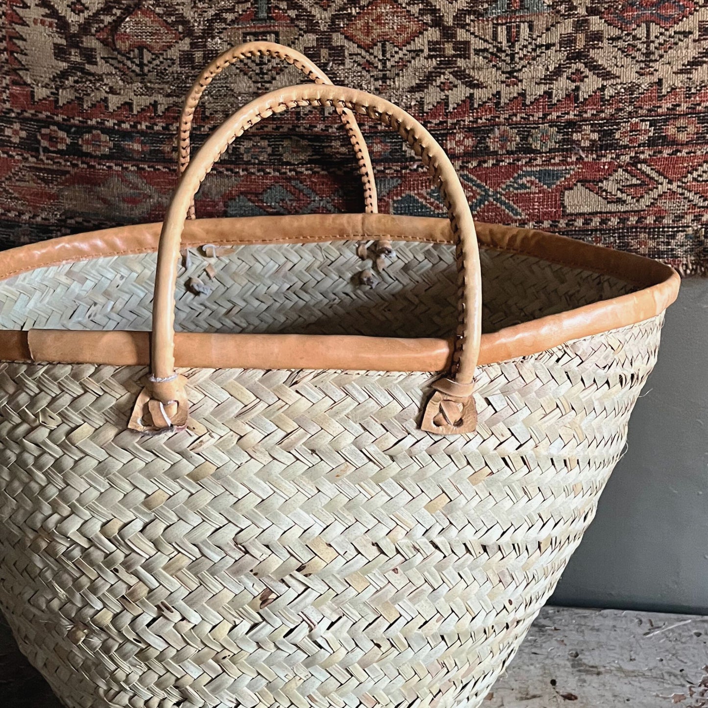 A woven palm fiber tote basket with tan leather handles and a rug behind it.
