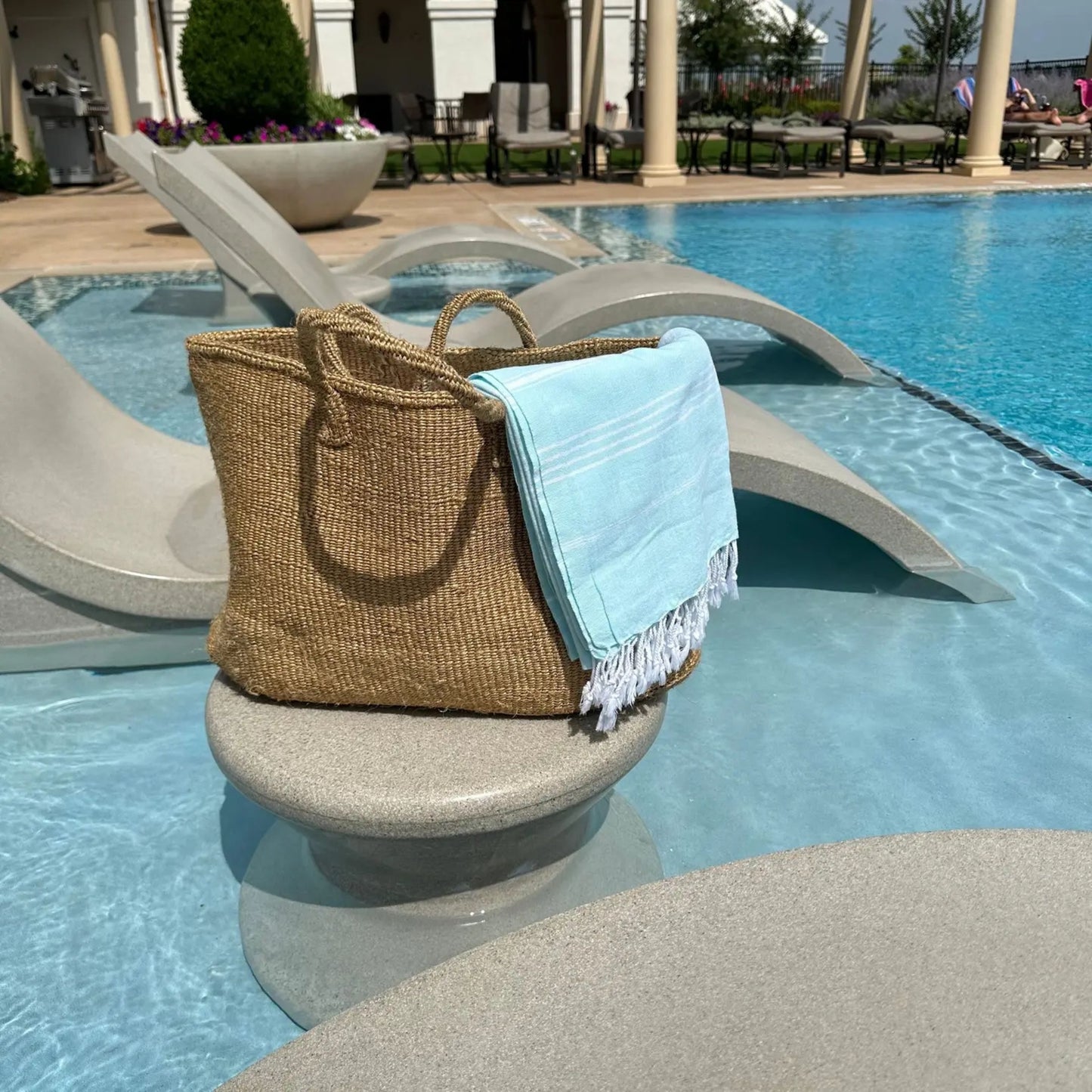 Woven bag with a blue towel on a stone ledge by a pool