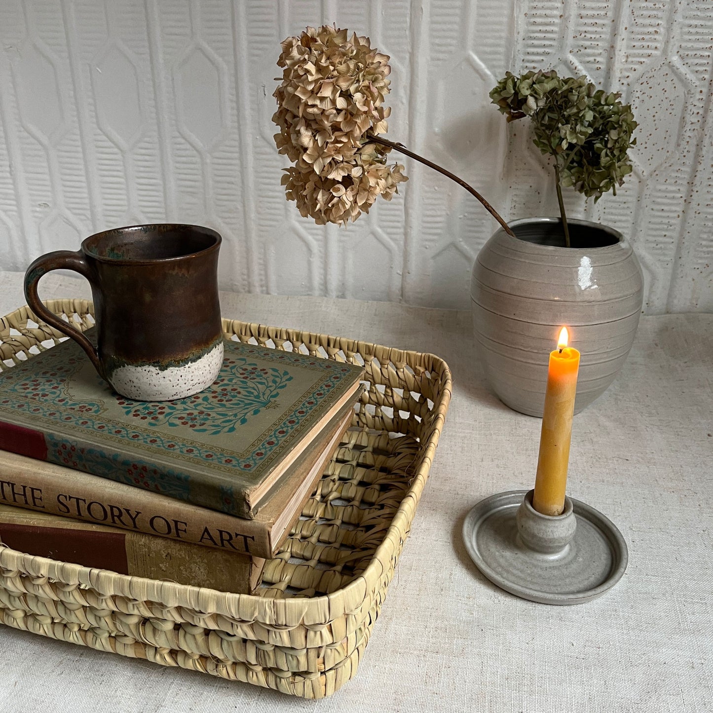 Decorative setting with a woven tray holding books and a mug, a candle in a holder, and a vase with flowers on a textured surface.