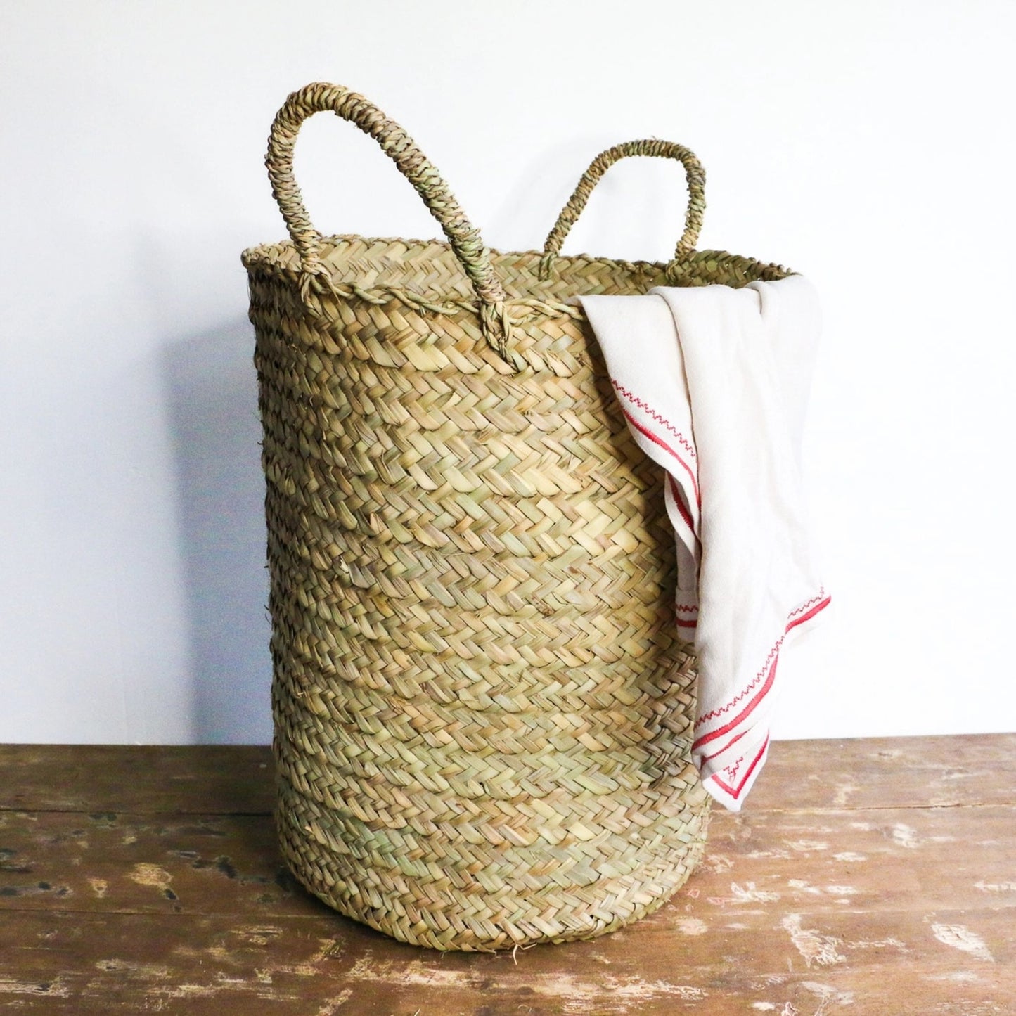 Woven hamper with handles on ground with white and red blanket hanging out of the top of it.