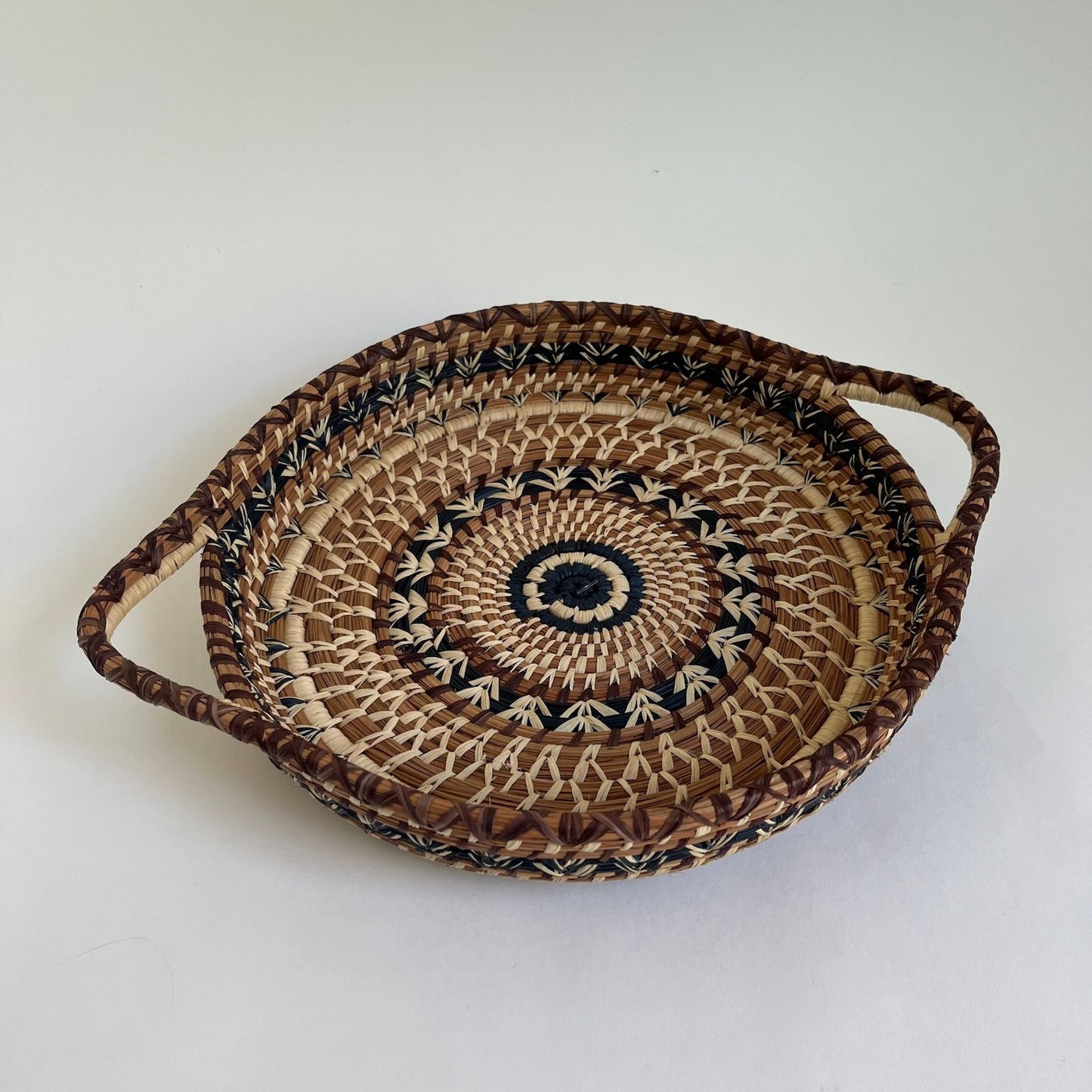 Shallow, round, pine needle tray basket with handles. It has a brown, black, and tan woven design on the top.