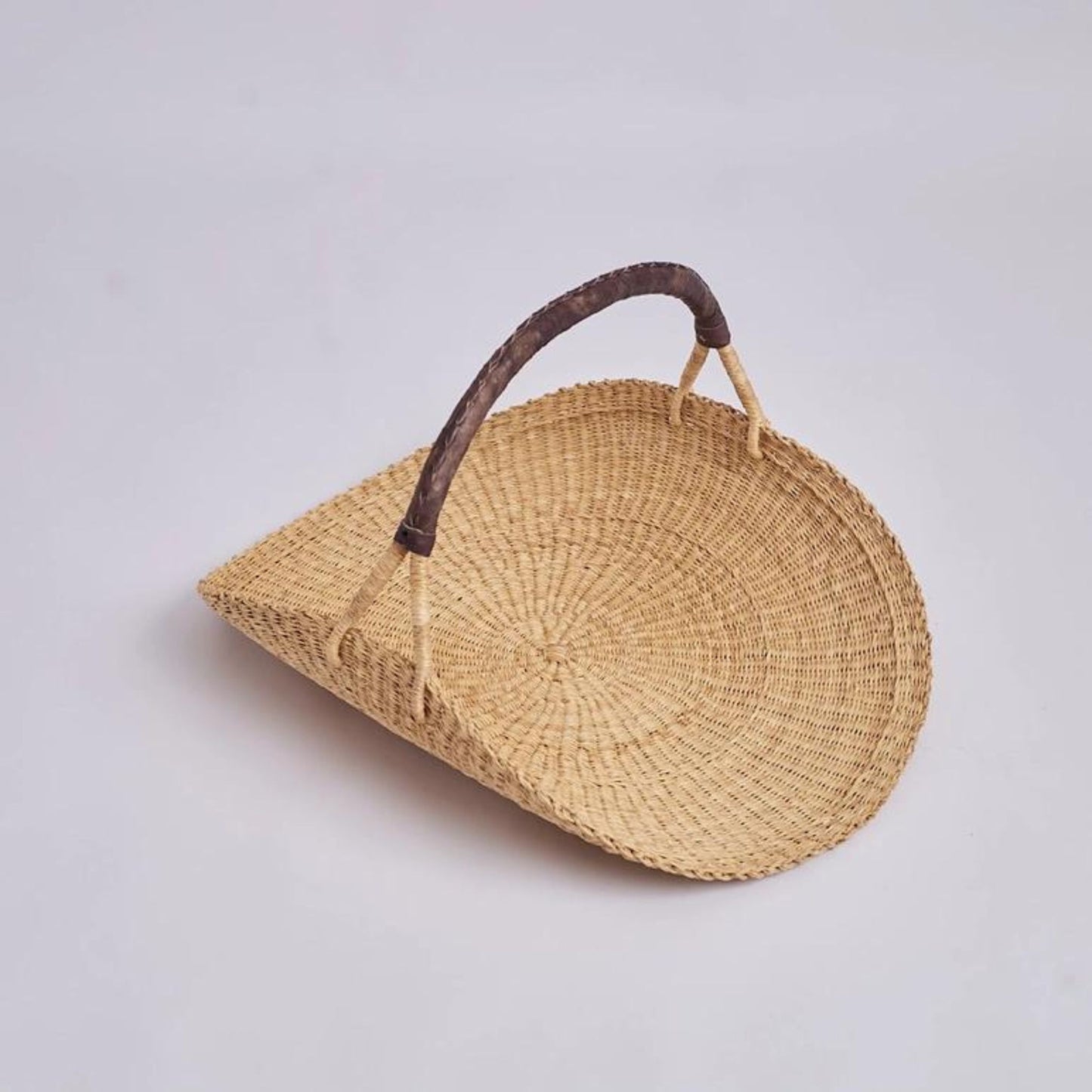 An oversized garden trug basket made from a flat woven disc that curves up at the brown leather handle.