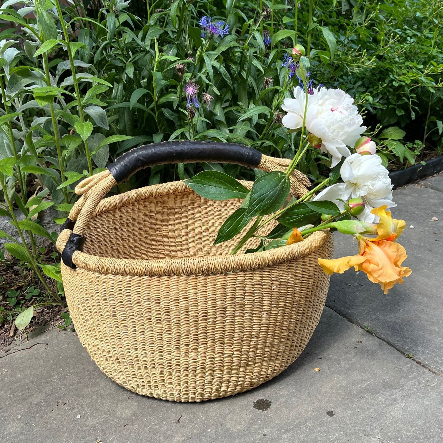 Large bolga basket with a black leather handle holding flowers outside.