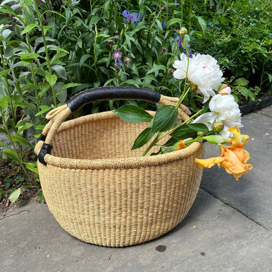 Large bolga basket with a black leather handle holding flowers outside.