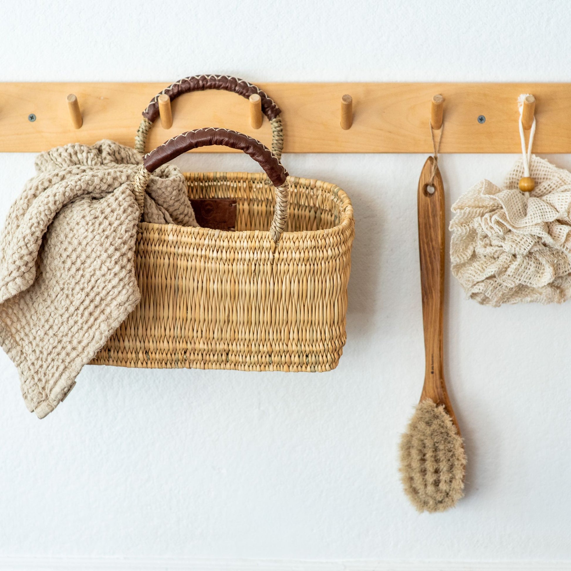 An oak bathing brush with natural fiber bristles hanging from a wall rack alongside a loofah and rectangle basket holding a waffle linen towel.