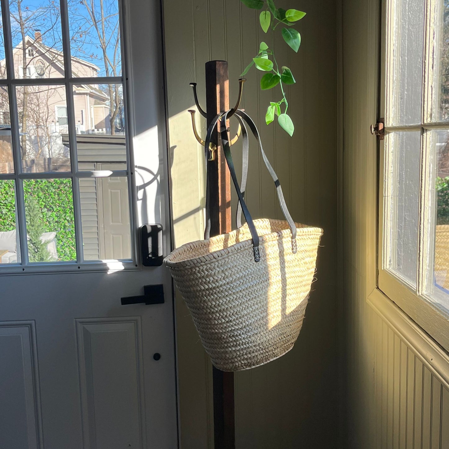 French market tote with black leather shoulder straps hanging from an entryway coat rack in between a door and window with a plant hanging from above.