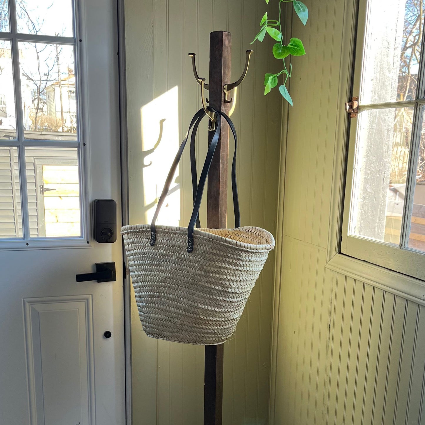 French market tote with black leather shoulder straps hanging from an entryway coat rack in between a door and window with a plant hanging from above.