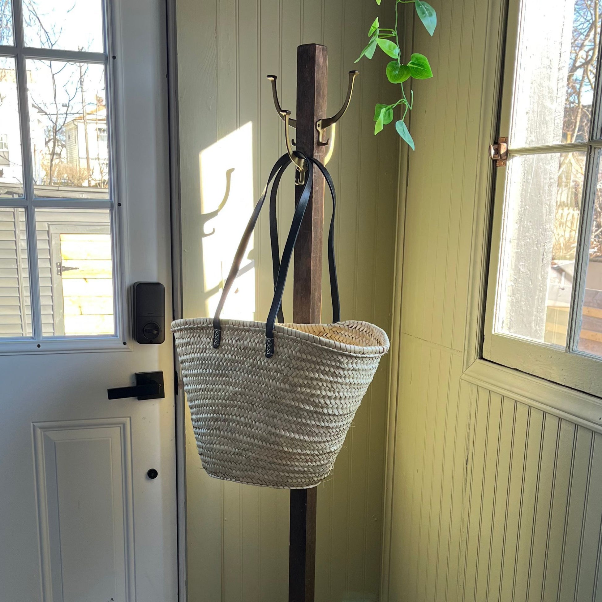 French market tote with black leather shoulder straps hanging from an entryway coat rack in between a door and window with a plant hanging from above.