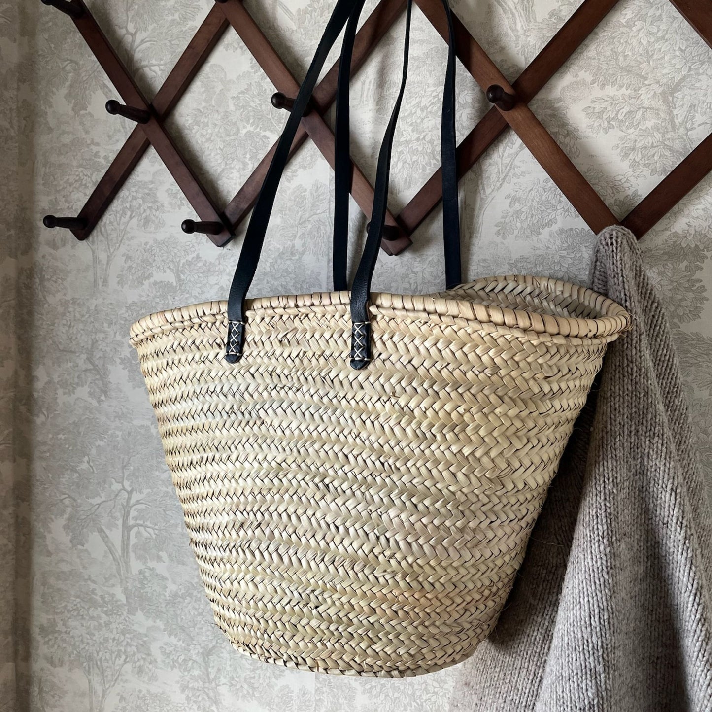 French market tote with black leather shoulder straps hanging from a hook on a wall alongside a grey sweater.