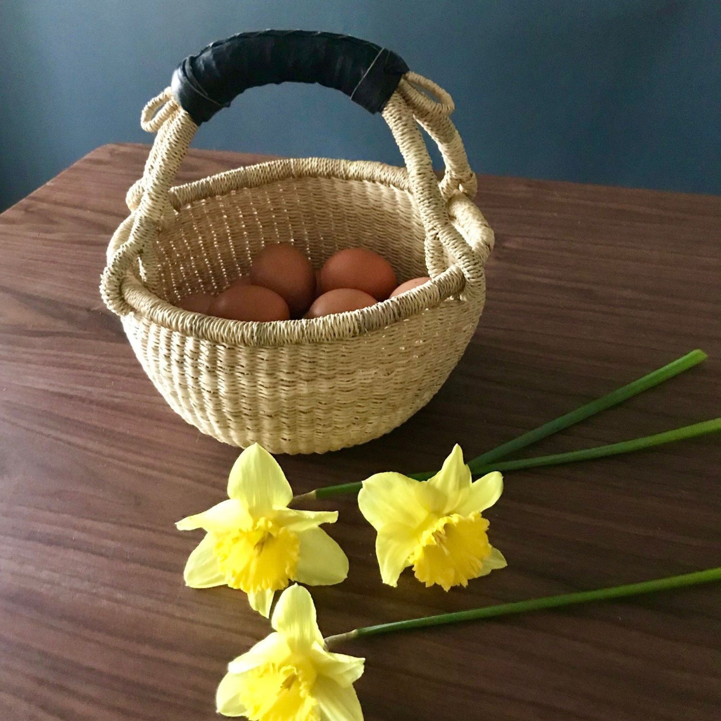 A mini round bolga basket with a black leather handle holding eggs next to daffodils.