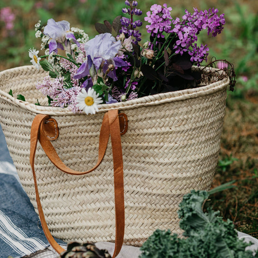 Woven tote basket with double leather shoulder strap and tote handles filled with flowers in the grass.
