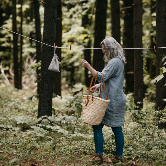 A woman holding a woven tote basket with double leather straps going to use a clothesline in the woods.