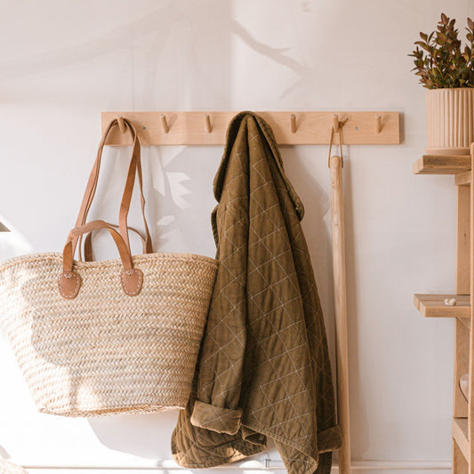 A woven tote basket with double leather shoulder strap and tote handle hanging on a wall hook next to a jacket and broom.