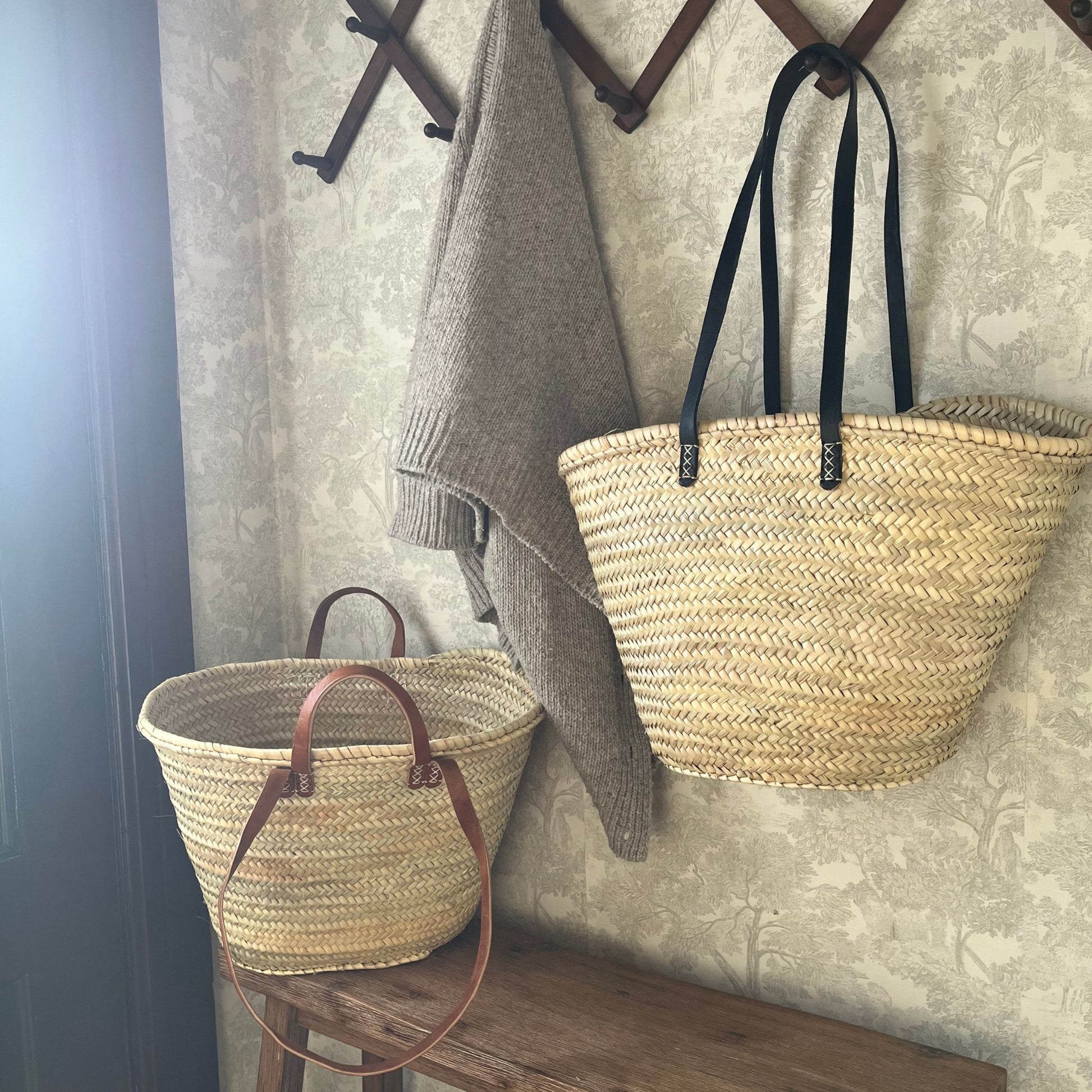 French market tote with black leather shoulder straps hanging from a wall hook alongside a grey sweater and a french market tote with tan straps sitting on a wooden bench indoors.