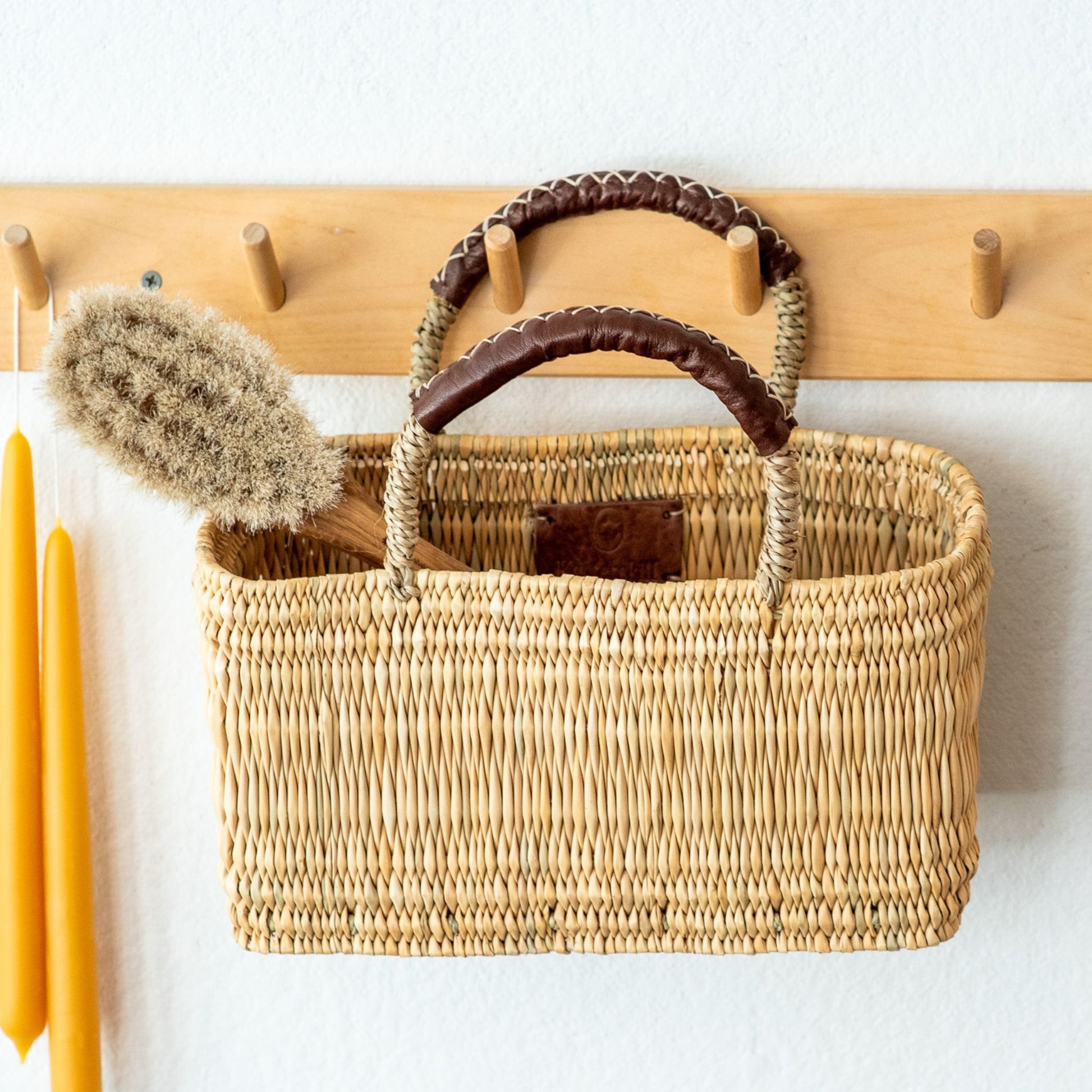 An oak bathing brush with natural fiber bristles inside a rectangle basket hanging from a wall rack alongside a pair of beeswax candles