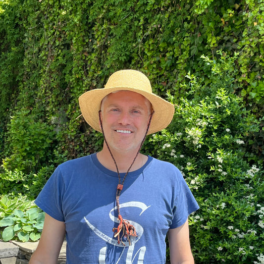A man wearing a tan, short brim straw sun hat with a braided straps outside with greenery in the background.