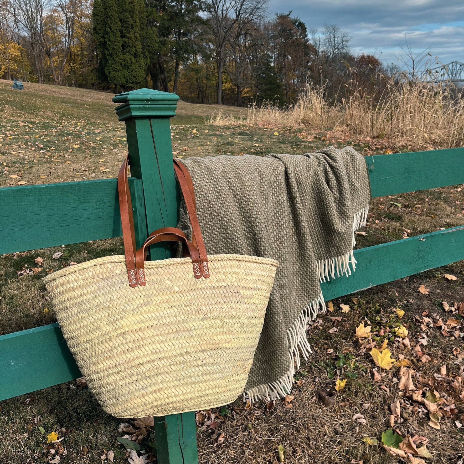 A french market tote bag with leather straps hanging from a fence next to a blanket draped over the fence.