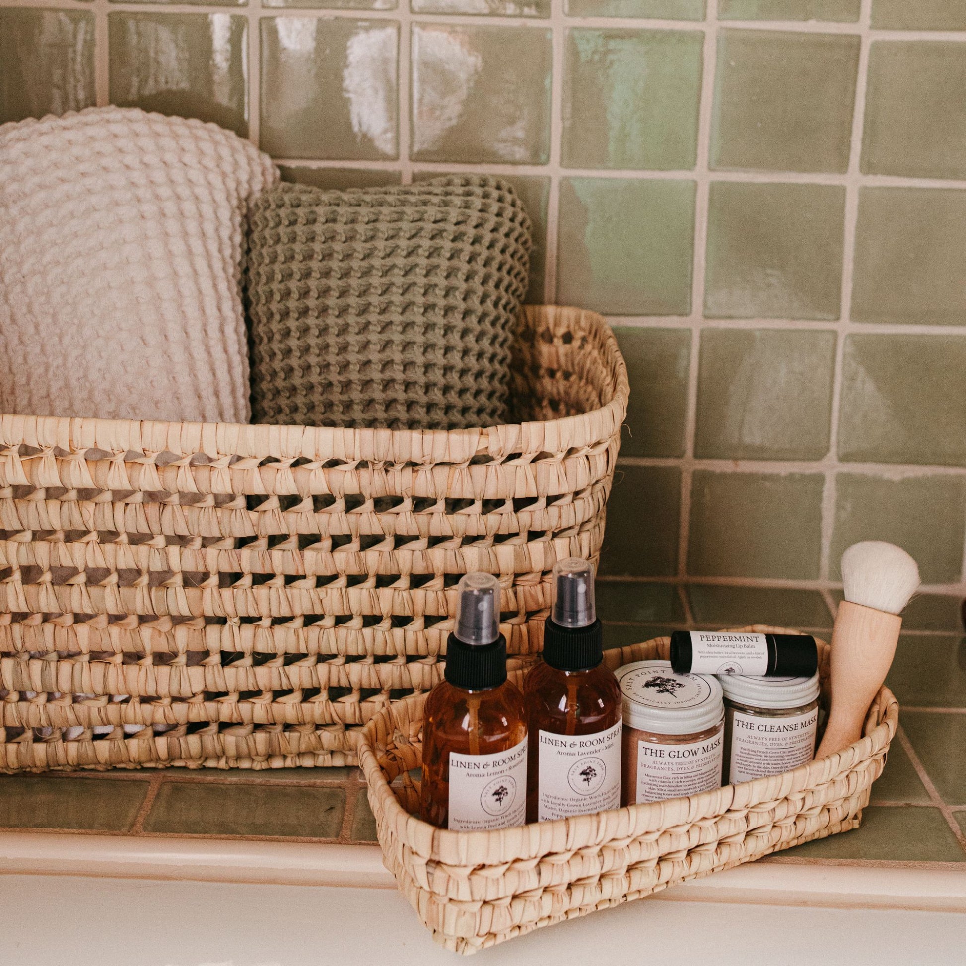 The Kabuki makeup brush alongside self-care products in a small basket alongside a larger basket holding linen towels all in a green tiled bathroom.