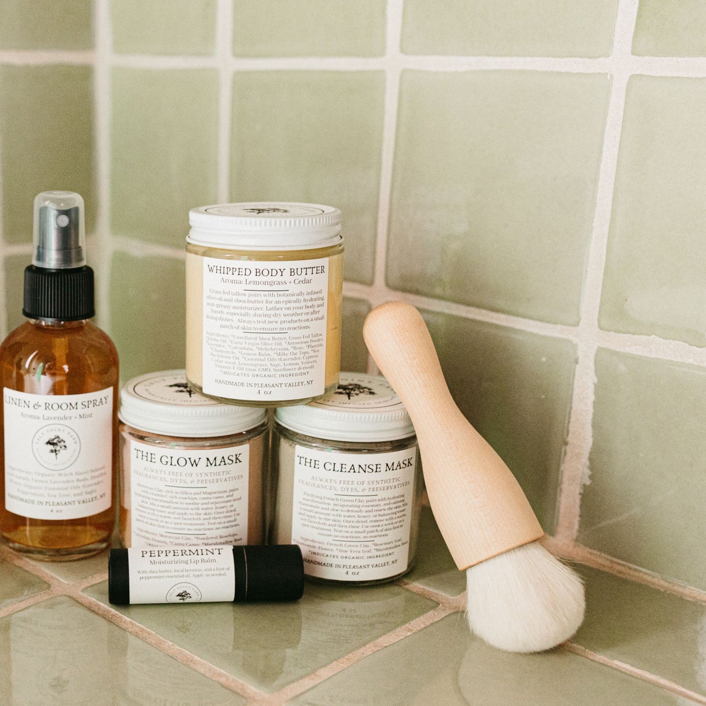 The kabuki makeup brush alongside face masks, lip balm. room spray, and body butter in a green tiled bathroon.