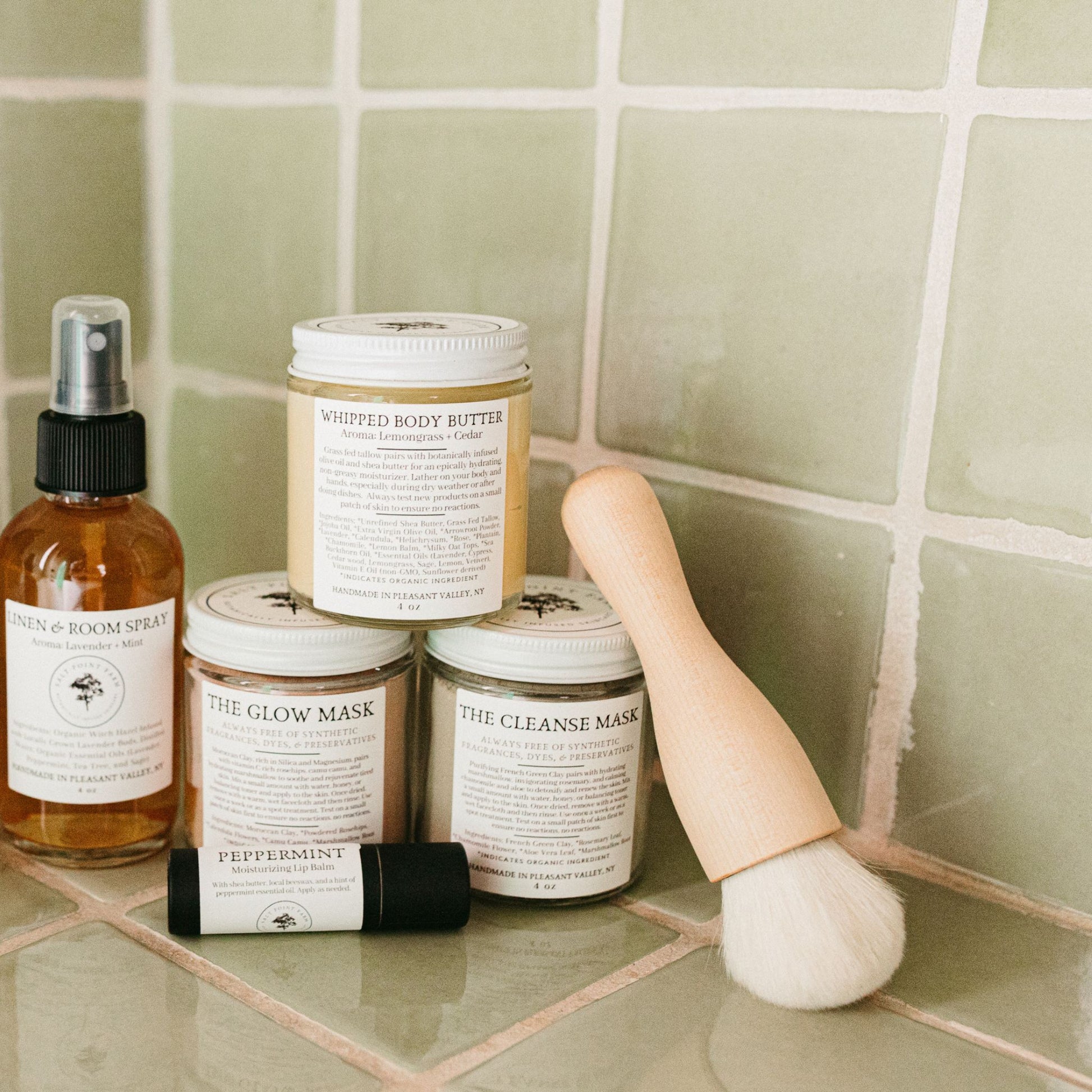 The kabuki makeup brush alongside face masks, lip balm. room spray, and body butter in a green tiled bathroon.