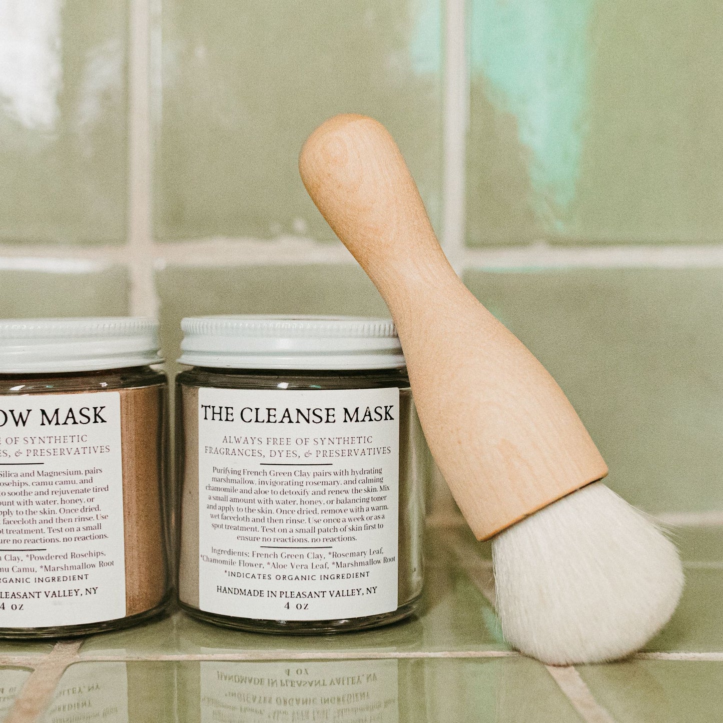 A goat hair and oiled birch wood makeup brush, alongside jars labeled 'THE CLEANSE MASK' and 'THE CLEANSING Balm' all in a green tiled bathroom.