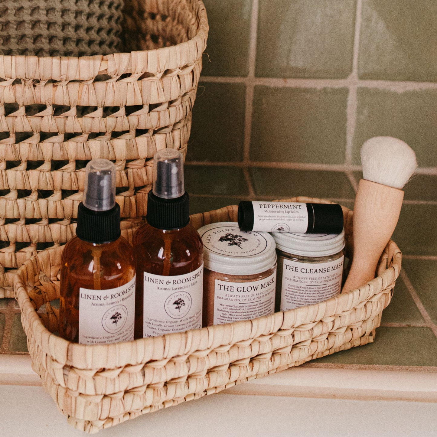 The Kabuki makeup brush alongside self-care products in a small basket in a green tiled bathroom.