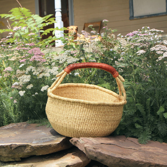 A natural woven straw basket with a curved reddish-brown wooden handle, resting on rustic stones in a garden setting with flowers and a house porch visible in the background.