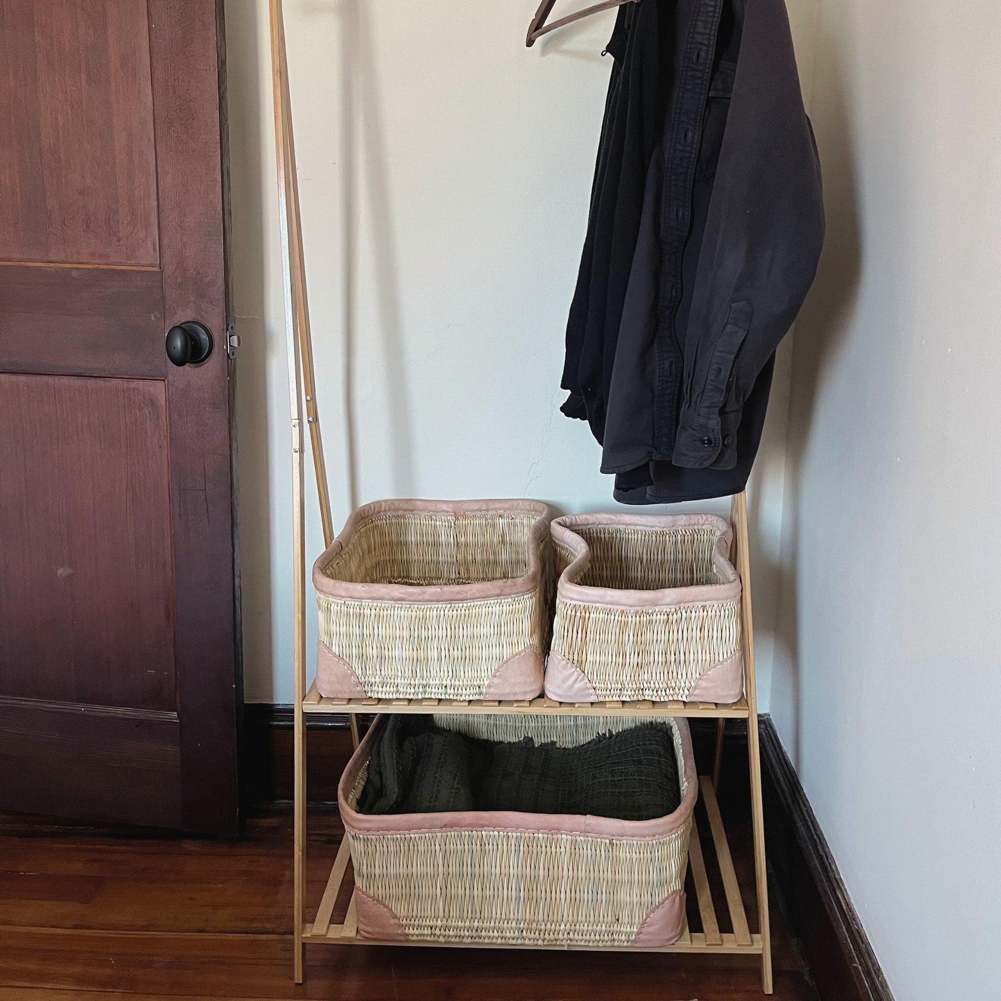 Three rectangular leather-trimmed woven storage baskets on a shelf being used to hold clothing in a closet.