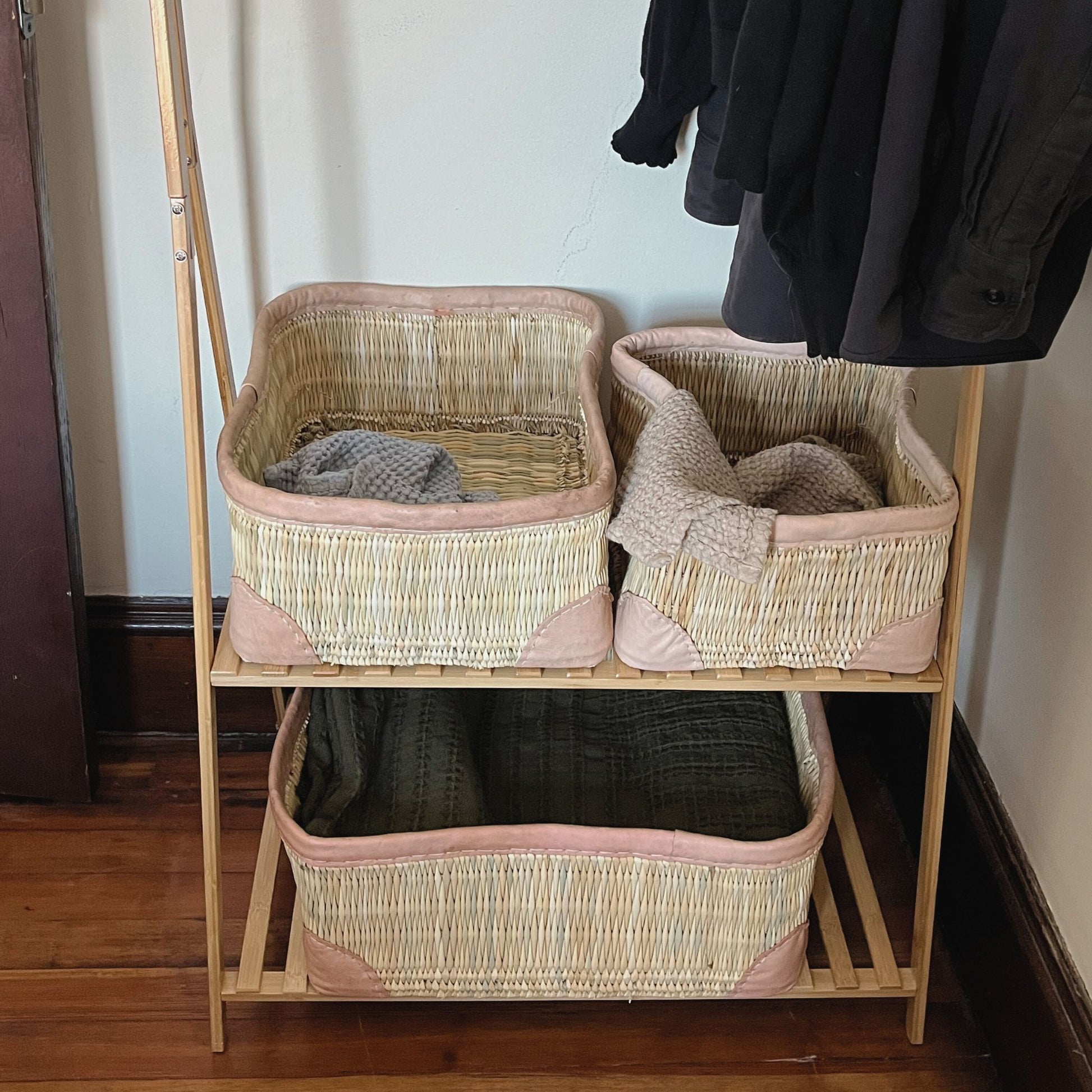 Three rectangular leather-trimmed woven storage baskets on a shelf being used to hold towels and a blanket.