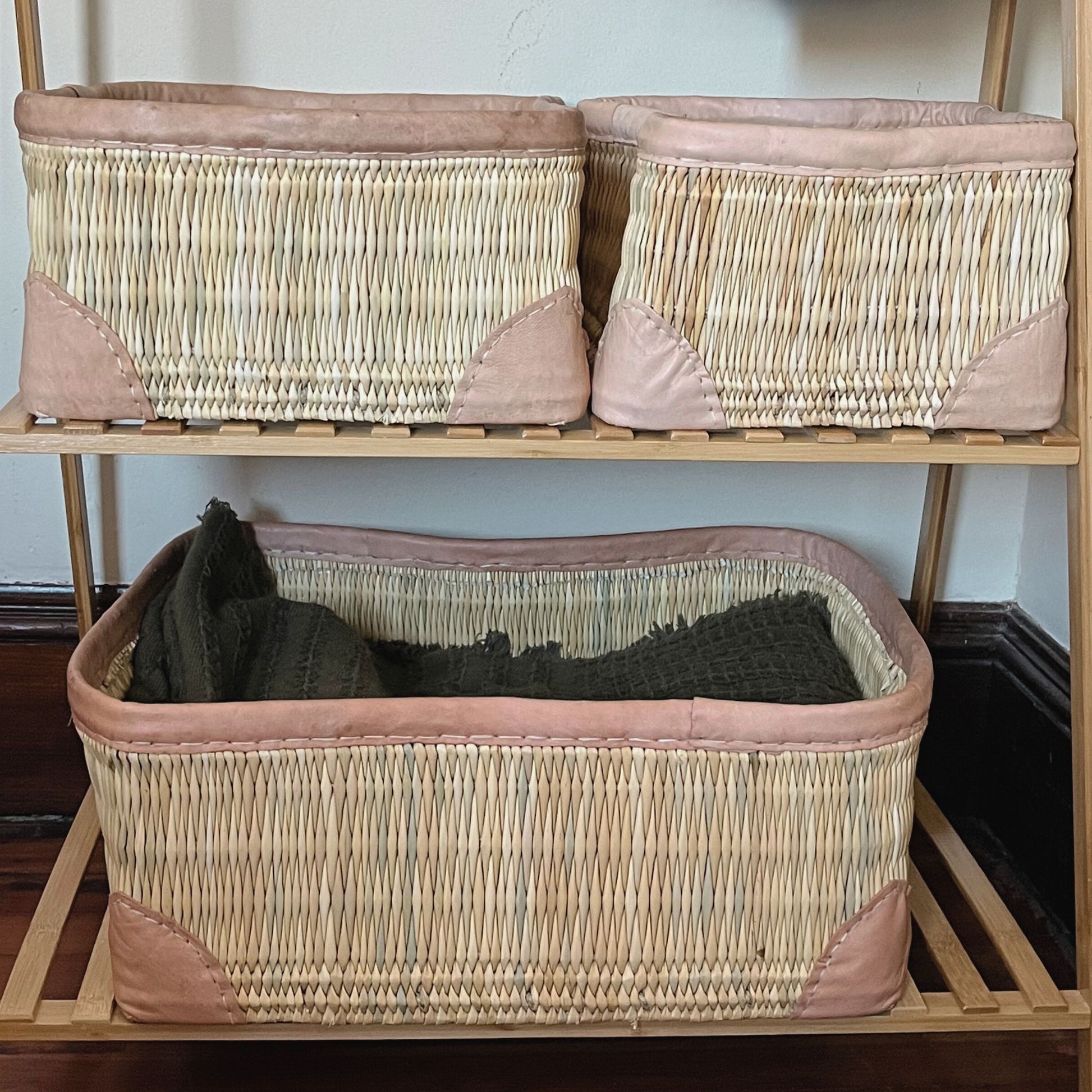 Three rectangular leather-trimmed woven storage baskets on a shelf being used to hold blankets.
